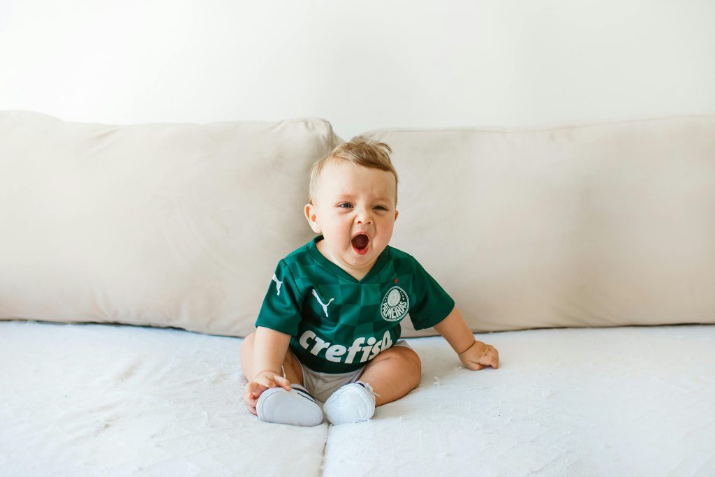 Cute baby wearing a green jersey yawning on a soft beige couch indoors.